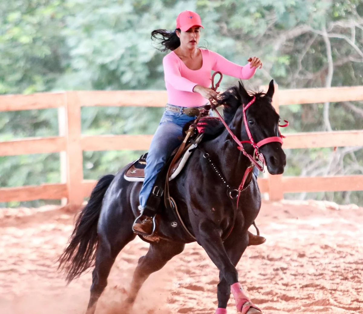 Dra. Mallu Cerutti a galope num cavalo preto na pista de areia, de boné e camisa rosa